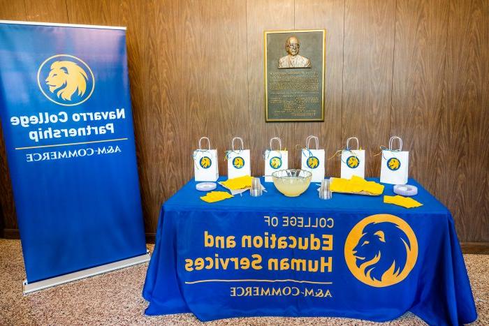 Goody bags and refreshments sit atop a table adorned with a blue and gold tablecloth next to an A&移动商务纳瓦罗学院伙伴关系旗帜横幅.